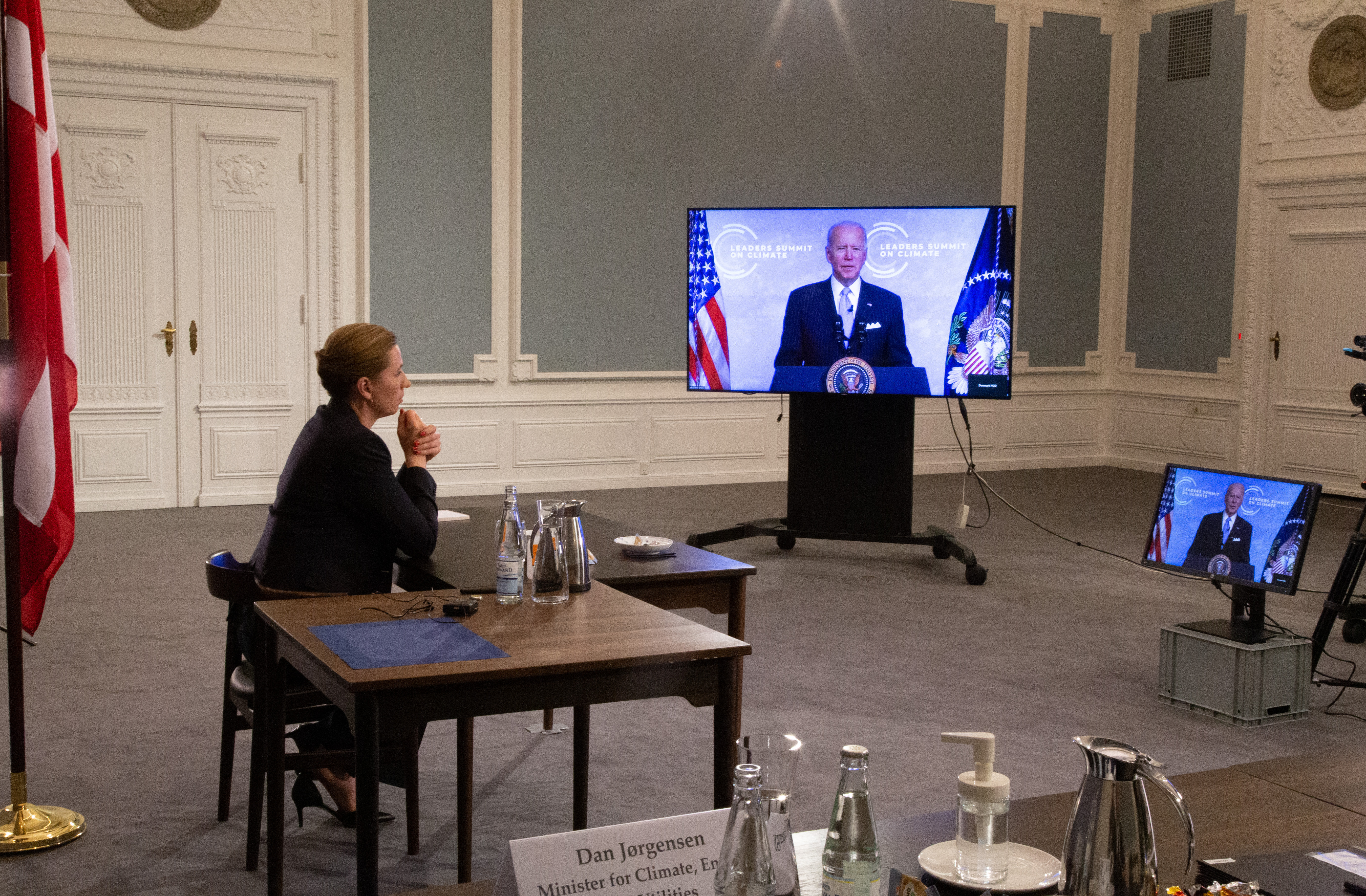 Mette Frederiksen sidder og holder virtuel konference. Hun sidder ved et bord og kigger hen på en fladskærm
