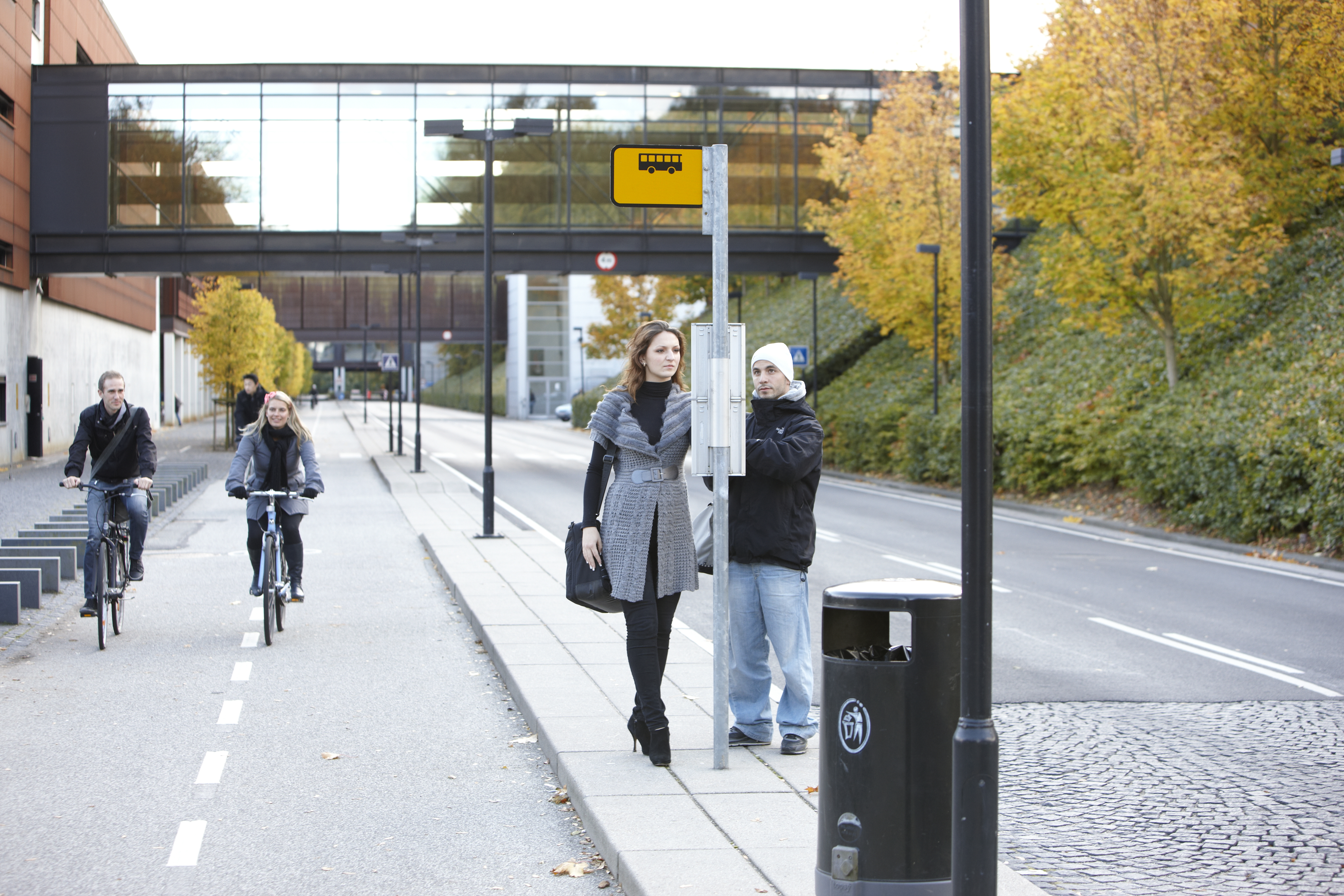 Cyklister kører forbi busstoppested med ventende passagerer foran Syddansk Universitet (SDU) i Odense.