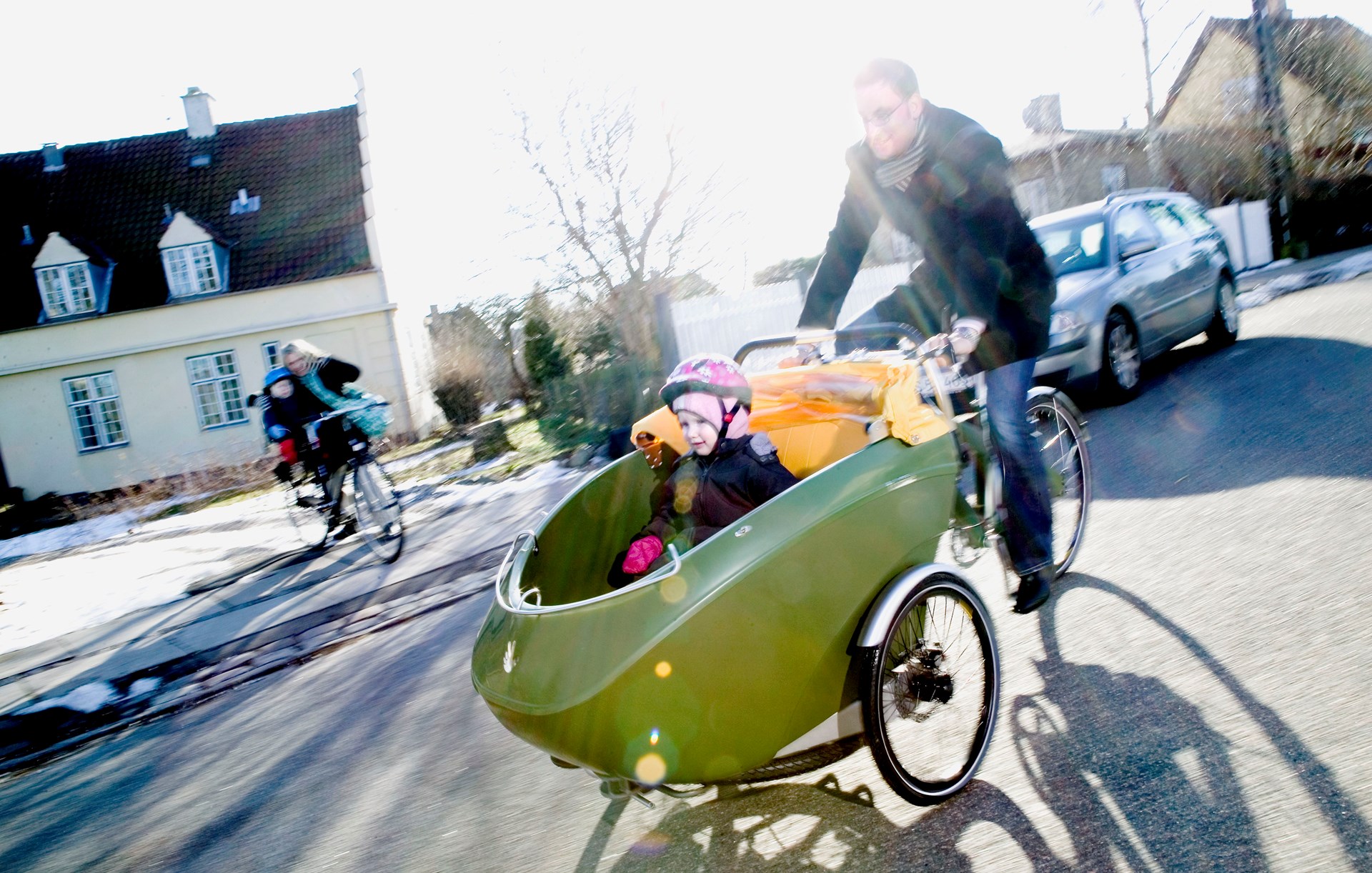 En mand cykler på en ladcykel. Heri sidder en pige med lyserød cykelhjelm