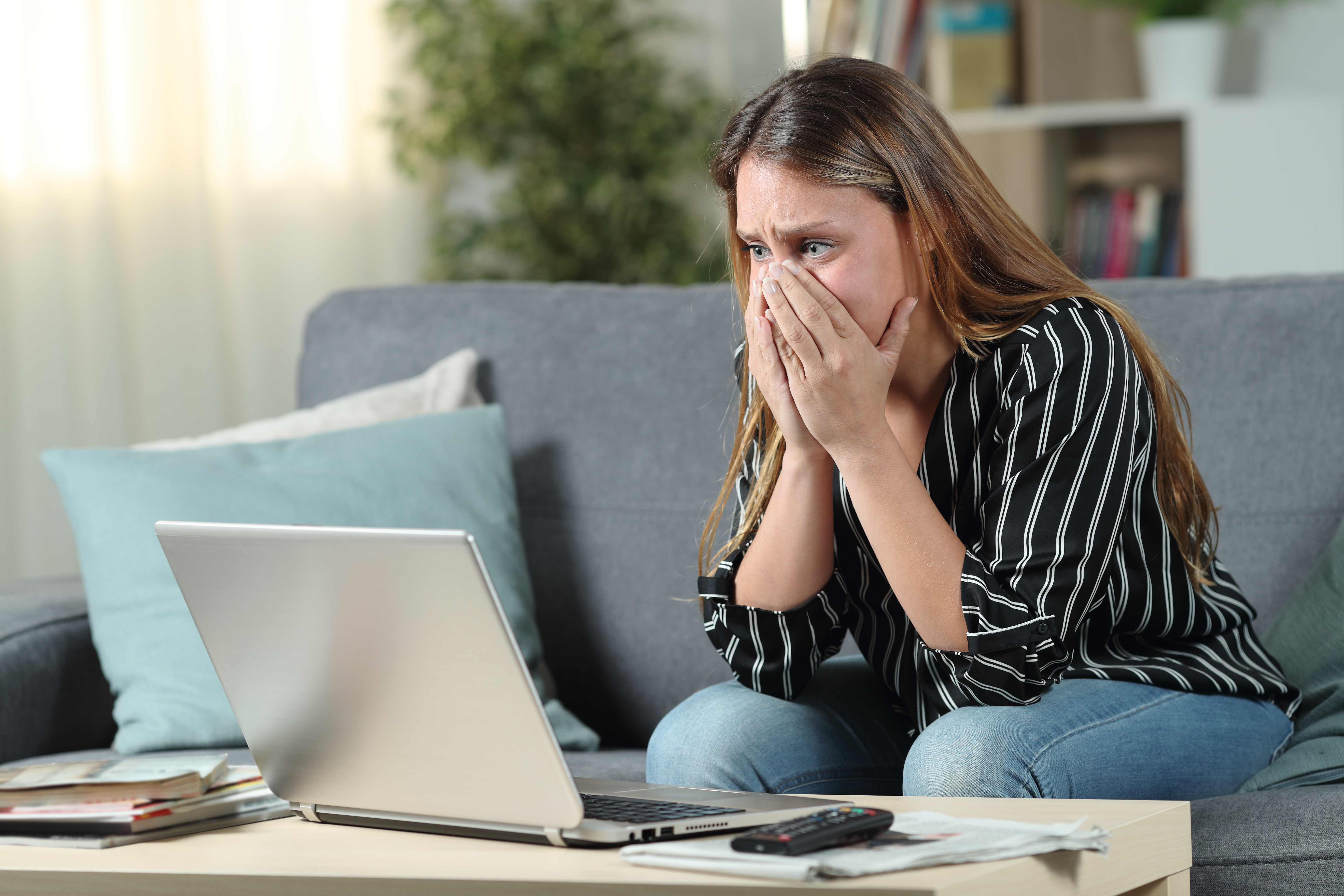 Kvinde sidder i sofa med hænderne op foran munden. Hun kigger ned på en bærbar computer, som står på et sofabord